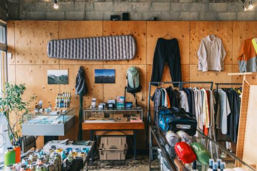 沖縄・宜野湾のアウトドアショップ「narrative outdoor supply」<br /><br /><br /><br /> 下書き保存<br /><br /> 公開<br /><br /> 31/100<br /><br /> 82<br /> /100<br /><br /><br /><br /> ブロックタイプまたはスタイルを変更<br /><br /> 画像ブロックを37から36へ上方向に移動<br /> 画像ブロックを37から38へ下方向に移動<br /> 配置を変更<br /><br /><br /> 【時松執筆中】海だけじゃない、沖縄の外遊びの新しい入口。宜野湾「Narrative Outdoor Supply」が作るローカルな提案。<br /> 米軍基地と沖縄の海がのぞめる宜野湾の通り沿いに、アウトドアギアやウエアを扱うショップ「Narrative Outdoor Supply（ナラティブアウトドアサプライ）」は店を構えている。　<br /><br /> 店内に並ぶのは、キャンプ用品やハイクギア、沖縄の街にも馴染むオリジナルウエアまで。アウトドアショップではあるけれど、いわゆる山道具の店とも、キャンプ道具だけの店とも少し違う。外遊びの道具と、グラフィック、ローカルなカルチャーが混ざった空間だ。<br /><br /> 沖縄のアウトドアといえば、真っ先に海を思い浮かべる人も多いはず。けれどナラティブが提案するのは、それだけではない。もっと身近に、もっと自由に外へ出て、自分なりの楽しみ方を見つける。沖縄の外遊びに、新しい入口をつくろうとしている店だ。<br /><br /> ▶︎画像1 キャプション案<br /> 米軍基地と海をのぞむ宜野湾の通り沿いにある「Narrative Outdoor Supply」。外遊びの道具とローカルカルチャーが混ざるショップだ。<br /><br /> 焚き火から始まった、「アクティビティからメディテーションへ」という考え方。<br /> 沖縄・宜野湾のアウトドアショップ「narrative outdoor supply」オーナー<br /> 代表の眞謝さん。グラフィックやプリントの制作活動も、店のオリジナルアイテムにつながっている。<br /> 現在ナラティブを代表として運営するのは、眞謝さん。ショップオーナーとして意外にも「MASH UP INK」というシルクスクリーンプリント工房を営み、「TOZOW MAJA / トゾウ マジャ」名義で個展も開催するグラフィックアーティストの顔を持つ。<br /><br /> アウトドアとの距離が近づいたのは、コロナ禍にキャンプ、とくに焚き火にハマったことがきっかけ。火を見ていると、頭のなかのざわつきが静まり、自分が何に惹かれているのか、自然と見えるようになってくる。<br /><br /> そんなキャンプ熱をSNSで発信していたところ、狩猟などにも親しむ共同創業者の大野さんと意気投合し、ナラティブを立ち上げるに至った。山やトレッキングに足を踏み入れるようになったのも、大野さんに誘われて里山へ入ったことがきっかけだったという。<br /><br /> 那覇でもやんばるでもなく、宜野湾にある理由。<br /> 沖縄・宜野湾のアウトドアショップ「narrative outdoor supply」<br /> アウトドアショップの立地として考えるなら、人が集まる街中か、フィールドに近い場所が多いだろう。沖縄でいえば那覇や、やんばるのような北部エリアだ。<br /><br /> けれど、ナラティブが店を構えたのは、沖縄県中部の宜野湾だった。<br /><br /> 理由のひとつは、宜野湾が「街と自然の中間地点」にあること。那覇方面からも訪れやすく、北部の自然豊かなフィールドにも足を延ばしやすい。眞謝さん自身の活動拠点がもともと宜野湾だったこともあり、ここが現実的で動きやすい場所だったという。<br /><br /> 沖縄・宜野湾のアウトドアショップ「narrative outdoor supply」オーナー<br /> そして、この立地は、店が提案したい外遊びともつながっている。最近始めているのが、店から読谷方面まで歩き、ビーチでテント泊をして戻ってくるような「シティハイク」だ。<br /><br /> 沖縄は車移動が当たり前で、長い距離を歩くことが文化としてあまり根付いてはおらず、珍しく見える土地でもある。だからこそ、あえて街を歩く。途中で飲食店に立ち寄ったり、写真を撮ったりしながら歩いて、ビーチでテントを張る。観光でも登山でもない、沖縄の新しい遊び方がそこにはある。<br /><br /> ▶︎画像3 キャプション案<br /> 宜野湾は、街にも自然にもアクセスしやすい中間地点。店の提案する外遊びとも相性がいい。<br /><br /> 古いコインランドリーに、アウトドアギアと工房を詰め込んで。<br /> 沖縄・宜野湾のアウトドアショップ「narrative outdoor supply」オーナー<br /> 古い建物の質感を活かした店内。木の什器が、アウトドアギアの並びに温かさを加えている。<br /> 店舗が入る建物は、もともとコインランドリーだった場所。天井は高く、ところどころに古い建物らしい荒さも残っている。<br /><br /> 内装で目を引くのは、三角形の什器や壁面に使われた針葉樹合板。眞謝のイメージしたものを、先輩の家具職人にワンオフで作ってもらったものだという。古い建物の無機質さに、木の温かさが加わって、店内には独特の落ち着きがある。<br /><br /> 沖縄・宜野湾のアウトドアショップ「narrative outdoor supply」オーナー<br /> 店の奥には「MASH UP INK」のプリント工房も併設。オリジナルアイテムを自分たちの手で作れるのも特徴だ。<br /> 店舗の奥には、眞謝さんが長年続けてきたシルクスクリーン工房「MASH UP INK」がある。今年からこの場所へ移転し、ナラティブの店内でプリント作業まで行えるようになった。<br /><br /> 元店長が独立して始めた刺繍の作業場も同じ空間にあり、キャップやウエアの刺繍、プリントまで、店の中で形にできるのがこの場所の強みだ。セレクトショップでありながら、自分たちで作ることもできる。ナラティブの空間には、売る場所と作る場所が地続きになっている面白さがある。<br /><br /> 外遊びを、メディテーションの時間へ。<br /> 沖縄・宜野湾のアウトドアショップ「narrative outdoor supply」オーナー<br /> 店名の「Narrative」には、自分だけの体験や物語を育てていくような意味が込められている。<br /><br /> 眞謝さんが掲げるのは、「アクティビティからメディテーションへ」という考え方だ。ここでいうメディテーションとは、特別な作法としての瞑想ではない。焚き火を眺めたり、山を歩いたり、目の前の自然や身体の動きに意識を向けることで、頭のなかにある余計な思考を少しずつほどいていく。そんな時間を指している。<br /><br /> ﻿<br /><br /> 自分の感覚がクリアになると、何に惹かれるのか、自分にとって何が心地いいのかも見えやすくなる。外で遊ぶことを通して、自分なりの基準を知る。その先で、道具の選び方や日々の過ごし方まで少しずつ変わっていく。<br /><br /> ナラティブが提案しているのは、ただアクティブに動くための外遊びではない。自然のなかで過ごす時間を、自分自身と向き合うきっかけにもしてほしい。そんな思いが、店名にも、店頭に並ぶ道具にも通っている。<br /><br /> 道具はシンプルに。目の前の自然を楽しめるセレクト。<br /> 画像<br /> 画像ファイルをアップロードするか、メディアライブラリから選択、または URL を追加してください。<br /> アップロード<br /> メディアライブラリ<br /> URL から挿入<br /> ナラティブのセレクトで眞謝さんが意識しているのは、シンプルで安心感があり、初めて使う人にも扱いやすいものだという。<br /><br /> それは、目の前の自然を楽しむために、道具の扱いで頭のキャパを使いすぎてほしくないから。使い方が複雑すぎず、フィールドで素直に頼れるもの。Rab（ラブ）やEXPED（エクスペド）など、ベーシックな海外ブランドのアイテムが多いのも、その考え方とつながっている。<br /><br /> ﻿<br /><br /> 一方で、沖縄ならではの気候や暮らし方もセレクトには表れている。近年は本土でも、アウトドアブランドのシェルが山道具としてだけでなく、街で着るファッションアイテムとして浸透してきた。<br /><br /> 車移動が多く、雨が降っても傘を差さない人が多い沖縄なら、雨をしのげて見た目もいいウインドシェルやレインウエアは、もっと日常に馴染むはず。眞謝さんは、そうしたアイテムが沖縄の街でも自然に選ばれるようになる可能性を感じているという。<br /><br /> ▶︎画像6 キャプション案<br /> RabやEXPEDなど、扱いやすさと信頼感のあるブランドも揃う。初めての道具選びにも入りやすい。<br /><br /> オリジナルもセレクトも、街と外遊びを行き来するものを。<br /> ﻿<br /><br /> ナラティブの面白さは、セレクトだけでなく、自分たちで作れることにもある。店の奥にはプリント工房があり、刺繍の作業場も隣接。オリジナルウエアやキャップには、グラフィックやディテールを自分たちの手で形にできる店ならではの強みが出ている。<br /><br /> 店頭でもまず目を引いたのは、オリジナルのセットアップやTシャツ類。一方で、店頭には外へ出る気分をアゲてくれる小物も並んでいた。<br /><br /> 沖縄の街とフィールドを行き来する、オリジナルの2WAYセットアップ。<br /> ﻿<br /><br /> オリジナルアイテムの中でも存在感があったのが、「Narrative Original / 2Way Shirts」と「Narrative Original / 2Way Guide Pants」。どちらも透湿性・簡易防水を備えたナイロン100％の生地を使い、街でも外遊びでも着やすいバランスに仕上げたウエアだ。<br /><br /> ﻿<br /><br /> シャツは、袖をファスナーで取り外せる2WAY仕様。長袖としても半袖としても着られるので、日差しの強い時期や、旅先での温度変化にも対応しやすい。フロントポケットをあえて排除し、背面には大容量のゲームポケットを配置。ザックを背負ったときの干渉を避けつつ、街歩きや移動中でも手ぶらで動きやすい作りになっている。<br /><br /> ﻿<br /><br /> パンツも同じく2WAY仕様で、左右の裾を取り外せばハーフパンツとして着用可能。ジップ付きポケットや裾のドローコードなど、移動中や外遊びで使いやすいディテールも備える。<br /><br /> セットアップで着れば、街、旅、フィールドをそのまま行き来できる。沖縄の気候や街の空気にも馴染みつつ、外で使うことまで考えられた、ナラティブらしいウエアだ。<br /><br /> ▶︎画像7 キャプション案<br /> 袖や裾を取り外せる、Narrative Outdoor Supplyの2WAYセットアップ。街にも外遊びにも馴染む一着。<br /><br /> ▶︎画像8 キャプション案<br /> ボディから作り込んだNarrative Outdoor Supplyのオリジナルキャップ。長めのツバと刺繍が印象的で、街歩きにも外遊びにも取り入れやすい。<br /><br /> リフレクターのグラフィックが効いた、Parallel Teeシリーズ。<br /> ﻿<br /><br /> ナラティブのオリジナルTシャツ「Parallel」シリーズもナラティブのアイコン的アイテム。グラフィックを主役にしながら、夜道やアクティビティでの視認性にも配慮されているのが特徴だ。<br /><br /> 長袖の「Parallel L/S T-shirts」は、光が当たると反射するリフレクティブプリントを採用。吸汗速乾性やUVカット機能を備えたポリエステル100％の生地で、タウンユースはもちろん、ラッシュガード的にも使いやすい。<br /><br /> ﻿<br /><br /> 半袖の「Parallel Tee 4th」は、4周年記念で作られたモデル。チャコールのボディにリフレクターインクを使ったグラフィックが入り、車のライトやカメラのフラッシュに反応する。プリント工房を持つナラティブらしさが出た一枚だ。<br /><br /> ﻿<br /><br /> なお、現在はオリジナルのバンダナスカーフも再生産中だという。沖縄のヴィンテージマップをベースに、タトゥーアーティストのKENTA氏がデザインを手がけた大判スカーフで、再入荷したらこちらもチェックしたい。<br /><br /> ▶︎画像9 キャプション案<br /> リフレクティブプリントを取り入れた「Parallel」シリーズ。グラフィックと機能を同じ温度で楽しめるTシャツだ。<br /><br /> ▶︎画像10 キャプション案<br /> 再生産中のオリジナルバンダナスカーフ。ヴィンテージの沖縄地図をベースにした、スーベニア感のある一枚。<br /><br /> ネオンカラーのパラコードが映える、PIQNICのボトルホルダー。<br /> ﻿<br /><br /> セレクトアイテムでは、PIQNIC（ピクニック）の「Bottle Holder Bag 001」も印象的だった。パラコードを編み込んだような立体感と、ネオンカラーの配色が目を引く、ハンドメイドのボトルホルダーだ。<br /><br /> カラビナ付きでギア感もあり、街歩きやフェス、ビーチへの外出にも持ち出しやすい。500mlのペットボトルから1.0Lのナルゲンボトルまで対応し、ストラップの長さやショルダーカバーも調整できる。<br /><br /> ﻿<br /><br /> 水分補給の道具をバッグの中にしまい込まず、すぐ手に取れる場所に置いておけるのが便利。実用的でありながら、アクセサリー感覚でも楽しめる。こういう小物が自然に並んでいるところにも、ナラティブのセレクトの幅が出ている。<br /><br /> キャプション案<br /> ネオンカラーのパラコードとカラビナが目を引く、PIQNICのボトルホルダー。500mlペットボトルから1.0Lナルゲンまで対応。<br /><br /> ファミリーキャンプから、沖縄の外遊びを広げる店へ。<br /> ﻿<br /><br /> オープン当初のナラティブは、ファミリーキャンプ向けのアイテムも多く扱っていたという。キャンプブームの空気もあり、沖縄で外遊びに興味を持つ人の入口として、間口を広げる意味もあったのだろう。<br /><br /> ﻿<br /><br /> ただ、現在は少しずつ方向性を見直しているそうだ。より軽く、コンパクトで、街歩きや低山トレッキング、旅先での外遊びにも馴染む道具へ。眞謝さんが今考えているのは、沖縄でアウトドアを楽しむ選択肢を、海やキャンプ以外にも広げていくような売り場づくりだ。<br /><br /> ﻿<br /><br /> それは、単に山岳系に振り切るということではない。沖縄で歩くこと、低山を楽しむこと、街から海へ向かうこと。車移動が当たり前の土地で、あえて自分の足で移動してみること。そうした体験に合う道具を、これからもっと増やしていきたいという。<br /><br /> ナラティブは、いままさに沖縄のアウトドアを別の角度から見せようとしている途中にある。<br /><br /> 小さなコミュニティが生まれるイベントを増やしていく。<br /> ﻿<br /><br /> 今後について聞くと、眞謝さんは「小さなコミュニティができるようなイベントを増やしていきたい」と話してくれた。<br /><br /> これまでにも、ビーチで音楽を流しながらキャンプを楽しむイベントなどを行ってきたという。けれど今は、もっとコンパクトで、参加した人同士が自然に話せるような場にも関心があるそうだ。<br /><br /> ﻿<br /><br /> シティハイクのように、店を出発して、途中で飲食店に立ち寄り、歩けるところまで歩く。疲れたら途中で離脱してもいい。そんなゆるやかな企画なら、外遊びに興味はあるけれど一歩踏み出せなかった人にも届きやすい。<br /><br /> 沖縄のアウトドアを、海だけでなく、街や低山、日常の中にも広げていく。その入口を店とイベントの両方で作ろうとしているのが、ナラティブの現在地だ。<br /><br /> 宜野湾の空気ごと味わえる、アメリカンダイナーな「Stickywich」。<br /> ﻿<br /><br /> 周辺のおすすめとして教えてもらったのが、ナラティブと同じ通り沿いにあるサンドイッチショップ「Stickywich（スティッキーウィッチ）」。店の近くからは海と空が抜ける景色も見えて、目の前にはキャンプフォスター。通りを歩いているだけでも、宜野湾らしいアメリカンな空気が感じられるエリアだ。<br /><br /> ﻿<br /><br /> 店構えもかなりポップ。パープルとピンクを基調にした外観に、ネオンやステンレスのカウンターが合わさって、どこかアメリカのダイナーのようなムードがある。店内には海外のお客さんの姿も多く、メニューだけでなく空間ごと楽しめる一軒だった。<br /><br /> ﻿<br /><br /> 今回は、定番の「フィリーチーズステーキサンドイッチ」をポテトセットでオーダーした。サイズは通常、ハーフ、3分の1から選べるので、初めてならハーフでも十分満足できる。<br /><br /> ﻿<br /><br /> サンドイッチは、薄切りの牛肉とチーズがたっぷり入った食べ応えのある一品。ポテトもつけると、ランチとしての満足感はかなり高い。ドリンクには、沖縄ではおなじみのA&Wのルートビアも選べる。味も空間も含めて、宜野湾らしい寄り道として覚えておきたい。<br /><br /> ▶︎画像12 キャプション案<br /> キャンプフォスターの向かいにある「Stickywich」。パープルとピンクの外観が目を引く、ポップなサンドイッチショップ。<br /><br /> ▶︎画像13 キャプション案<br /> ネオンやステンレスのカウンターが印象的な店内。アメリカンダイナーのような空気も楽しい。<br /><br /> ▶︎画像14 キャプション案<br /> 定番のフィリーチーズステーキサンドイッチ。ポテトとルートビアを合わせれば、アメリカンな満足感たっぷり。<br /><br /> 沖縄の外遊びに、もうひとつの入口を作る店。<br /> ナラティブは、沖縄でアウトドアを楽しむための選択肢を、少しずつ広げようとしている店だ。<br /><br /> 海へ行くことも、山を歩くことも、街から外へ出てみることも、どれかひとつに決める必要はない。自分にとって心地いい遊び方を見つけ、その先でまた新しい景色に出会う。そんなきっかけを、道具やウエア、イベントを通してつくろうとしている。<br /><br /> 沖縄でいつもと少し違う外遊びにチャレンジしたくなったら、宜野湾のナラティブが、その入口になってくれるはずだ。<br /><br /> パネルを切り替え: AIOSEO 設定<br /> SERP プレビュー<br /><br /><br /> Favicon<br /> GO OUT WEB<br /> https://web.goout.jp › fashion › 488292<br /> 【時松執筆中】海だけじゃない、沖縄の外遊びの新しい入口。宜野湾「Narrative Outdoor Supply」が作るローカルな提案。 | ...<br /> 米軍基地と沖縄の海がのぞめる宜野湾の通り沿いに、アウトドアギアやウエアを扱うショップ「Narrative Outdoor Supply（ナラティブアウトドアサプライ）」は店を構えている。　 店内に並ぶのは、キャンプ用品やハイクギア、沖縄の街にも馴染むオリジナルウエアまで。アウトドアショップではあるけれど、いわゆる山道具 ...<br /> news のタイトル<br /> 以下のタグをクリックして、タイトルに変数を挿入します。<br /> すべてのタグを表示 →<br /> ﻿<br /> News のタイトル<br /> ﻿ ﻿<br /> 区切り<br /> ﻿ ﻿<br /> サイトのタイトル<br /> ﻿ <br /><br /><br /> 推奨される最大文字数60字のうち81字<br /> メタディスクリプション<br /> 以下のタグをクリックして、変数をメタディスクリプションに挿入します。<br /> すべてのタグを表示 →<br /> ﻿﻿<br /> News の抜粋<br /> ﻿﻿ <br /><br /><br /> 推奨される最大文字数160字のうち6379字<br /> 基礎となるコンテンツ<br /> 基礎となるコンテンツとは、コンテンツ戦略の基礎となる、サイト上の最も重要で有益な記事やページを指します。AIOSEO では、基礎となるコンテンツを次の用途に使用しています:リンクアシスタントの内部リンク推奨もっと詳しく →<br /><br /> 基礎としてマーク<br /> 基礎となるコンテンツは PRO の機能です。もっと詳しく →<br /> フォーカスキーフレーズ<br /> フォーカスキーフレーズを追加する<br /> 追加のキーフレーズを取得<br /> 追加のキーフレーズ<br /> キーフレーズの追加を追加する<br /> ページ分析<br /> Meta description length<br /><br /> The meta description is over 160 characters.<br /><br /> Content length<br /><br /> This is far below the recommended minimum of words.<br /><br /> Internal links<br /><br /> You are linking to other resources on your website which is great.<br /><br /> External links<br /><br /> Great! You are linking to external resources.<br /><br /> パネルを切り替え: アコーディオン<br /> アコーディオン コンテンツの登録<br /> アコーディオン<br /> Add Row<br /> パネルを切り替え: 記事オプション<br /> 紹介枠<br /> アイテム名説明文画像<br /> Add Row<br /> 画像ギャラリー<br /> 画像選択<br /> Add Row<br /> ショッピングカテゴリー<br /><br /> 未選択<br /> パネルを切り替え: RSS関連<br /> Antenna開始日時<br /> 記入フォーマット: 2016-09-01 02:00:00<br /><br /> Antenna終了日時<br /> 記入フォーマット: 2016-09-01 02:00:00<br /><br /> citrusカテゴリ<br /><br /> ライフスタイル<br /> Gunosyで非表示<br /><br /> Gunosyカテゴリ<br /><br /> コラム<br /> LINEで非表示<br /><br /> LINEカテゴリ<br /><br /> ファッション<br /> Clicccarで非表示<br /><br /> 記事広告としてRSS登録<br /><br /> ニコニコニュース タグ<br /> 最大４つまで選択可<br /><br /> 政治・社会ビジネスネット・科学海外スポーツエンタメゲーム・アニメ雑学<br /> Yahooで非表示<br /><br /> パネルを切り替え: YARPP: 関連投稿<br /> 関連投稿:<br /><br /> 仏の名門と新鋭ブランドが再タッグ。ボディオブワーク × メフィストの第2弾コラボが登場。 (46.022) <br /> 今すぐに着たいロングスリーブTとパンツがリーズナブルに。アウトレットで見つける春アイテム。【買えるGO OUT】 (50.938) <br /> 沖縄の「Narrative outdoor supply」が1周年！ アニバーサリーグッズを多数リリース。 (116.02) <br /> 10アイヴァンの超重厚セルフレームに即完カラーver.が登場。艶消し漆黒に職人ワザ光る。 (46.022) <br /> ダイニーマ製のアウトドア風呂敷。話題の「Narative」が仕掛ける、ワンタン別注の斬新作。 (67.115) <br /> 50以上のブランドを取り扱うアウトドアコンセプトショップが、沖縄・宜野湾に誕生！ (67.115) <br /> すべての一致を実際に表示するかどうか、またどのように表示するかは、YARPP の表示オプションによって異なります。 投稿を更新すると、一致を変更する場合があります。<br /><br /> 設定オプション 再読み込み<br /><br /> パネルを切り替え: NextScripts: Social Networks Auto Poster - Post Options<br /> URL to use for links, attachments and %MYURL%: Reset all SNAP data Shorten URL Auto - Post URL will be used<br /> Autopost to .... [Select All Accounts] [Unselect All Accounts] [Expand All Accounts] [Collapse All Accounts] [Show Only Selected] [Show All Accounts]<br /> Facebook (Facebook #0)<br /> Page - GO OUT JPN [104426765880028]<br /> Message Format:<br /> %TITLE% - %SITENAME% %URL%<br /> Post Type:(What's the difference?)<br /> Text Post - just text message<br /> Image Post - big image with text message<br /> Text Post with "attached"blogpost<br /> URL to use: Auto - Post URL or globally defined URL will be used<br /> Time to post: [Add] Immediately [Change]<br /> Twitter (Twitter 20240301)<br /> news<br /> ブロック<br /><br /> 画像<br /> 画像を挿入し、視覚に訴えます。<br /><br /> スタイル<br /><br /> デフォルト<br /><br /> 角丸<br /> 設定<br /><br /> アスペクト比<br /><br /> 元のサイズ<br /> 幅<br /> 自動<br /> px<br /> 高さ<br /> 自動<br /> px<br /><br /> 高度な設定<br /> 選択したブロックをスキップ<br /> 公開パネルを開く<br /> news<br /> 画像<br /> 通知画像ファイルをアップロードするか、メディアライブラリから選択、または URL を追加してください。<br /> ファイルを選択ファイルを選択ファイルを選択ファイルを選択ファイルを選択ファイルを選択<br /> ダイアログを閉じる<br /> メディアの選択またはアップロード<br /> ファイルをアップロードメディアライブラリ<br /> 詳細を展開<br /> メディアを絞り込む日付で絞り込む<br /> すべての日付<br /> 検索<br /> メディア一覧<br /> 81件 (229732件中) のメディア項目を表示中<br /><br /> さらに読み込む<br /> 添付ファイルの詳細<br /><br /> DSCF6166.jpg<br /> 2026年6月8日<br /> 1 MB<br /> 1200 x 800 ピクセル<br /> 画像を編集<br /> 完全に削除する Post Thumbnail Editor<br /> 代替テキスト<br /> 沖縄・宜野湾のアウトドアショップ「narrative outdoor supply」オーナー<br /> 画像の目的を説明する方法について、詳しくはこちらをご覧ください(新しいタブで開く)。装飾のみが目的の画像であれば、空欄にしてください。タイトル<br /> DSCF6166<br /> キャプション<br /> 説明<br /> ファイルの URL:<br /> https://web.goout.jp/app/uploads/2026/06/DSCF6166.jpg<br /> URL をクリップボードにコピー<br /> 選択されたメディアアクション選択<br /> 選択されていません