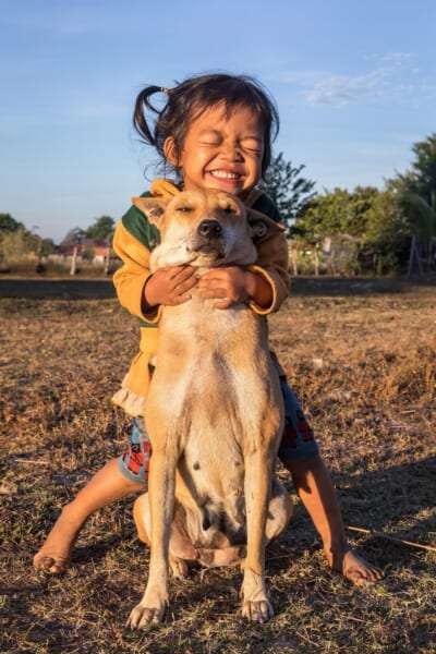 日の低い時間帯のやわらかな光の中で、2歳の女の子が笑顔で目を閉じ、自分の犬をぎゅっと抱きしめています(ラオス)。 人と犬という、種をこえた絆を写した一枚です。