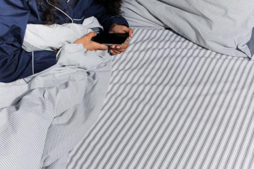 ベッドの上で、スマートフォンを両手で持つ人です(イメージ写真)。SNSを予定より長く使ってしまうかどうかも、のめり込み具合を測る質問の1つでした。